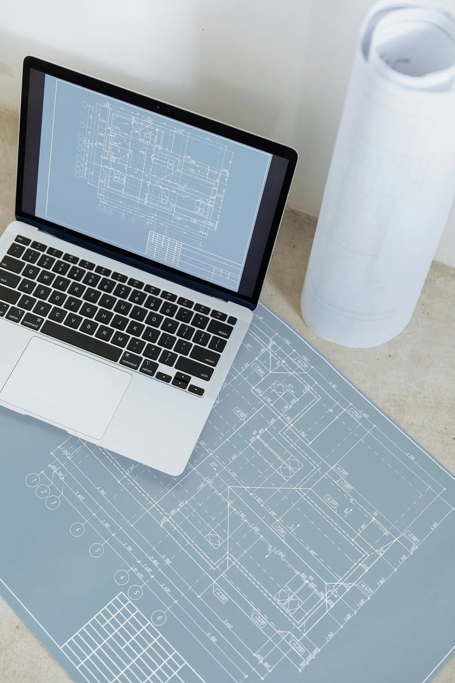 A laptop with architectural blueprints on the screen, placed on a table with more blueprints and a large water bottle nearby.