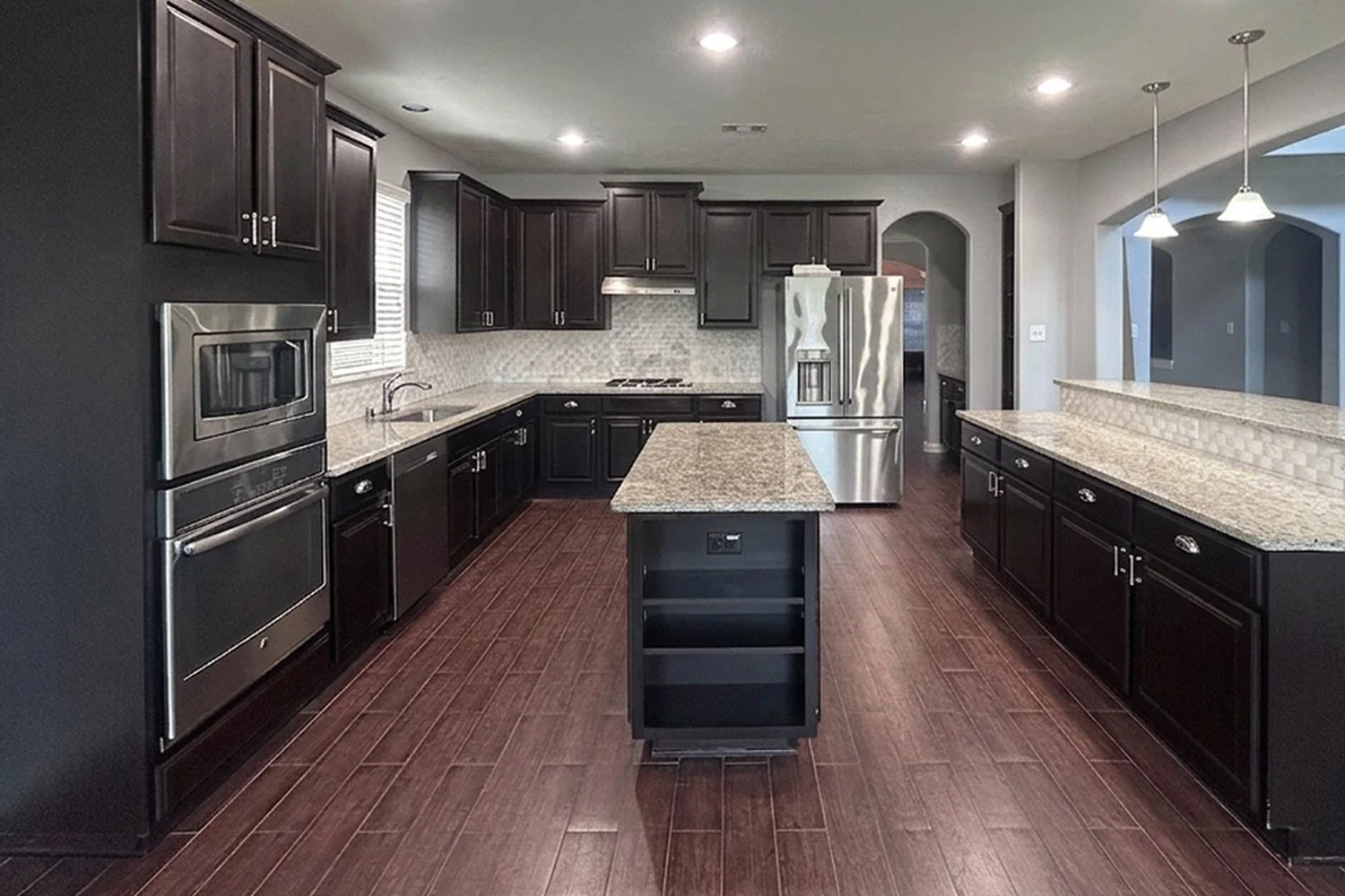 An outdated kitchen with dark cabinetry, stainless steel appliances, granite countertops, a center island, and hardwood flooring.