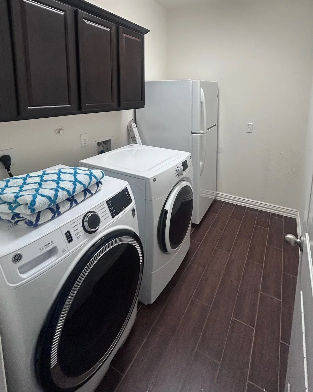 A dated, builder grade laundry room with dark wood cabinets, a washer and dryer, a white refrigerator, and dark wood floors.