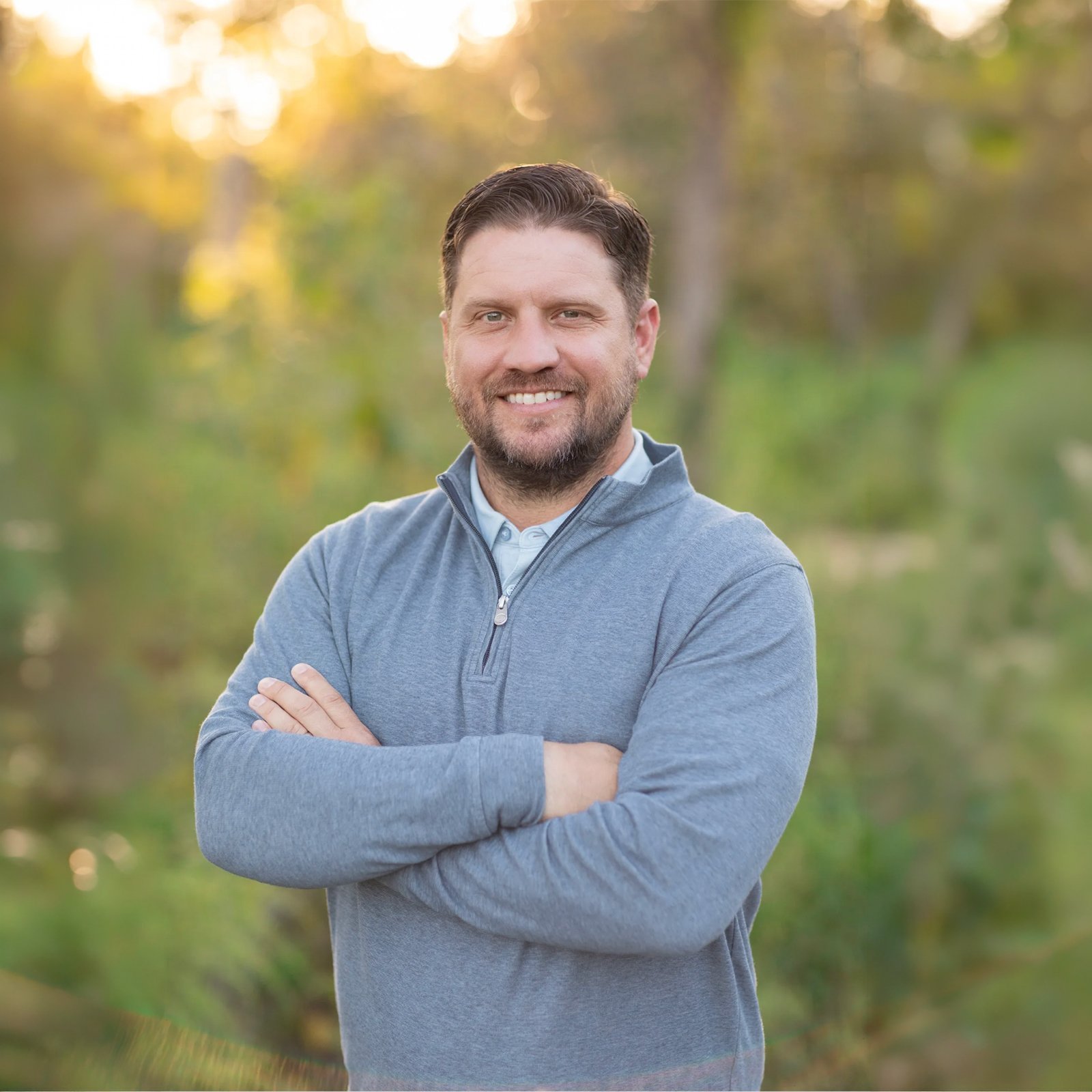 Mike Lehmann shown with brown hair and a beard, smiling, arms crossed, standing outdoors in a wooded area during sunset.