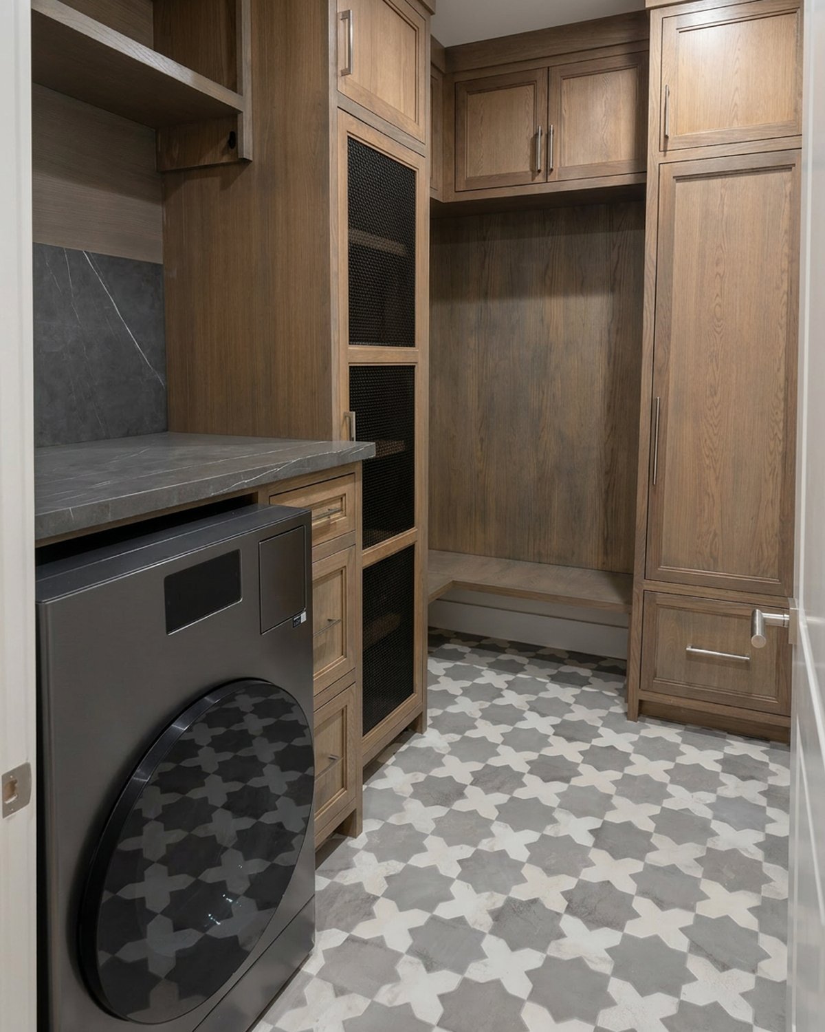 A modern, remodeled laundry room with custom wood cabinets, a modern washing machine, stone countertop, and patterned tile floor.