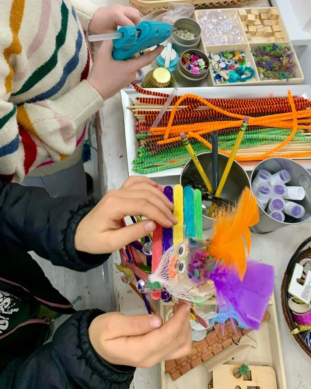 Children working on arts and crafts project with colorful pipe cleaners, pompoms, googly eyes, feathers, and markers on a cluttered table.