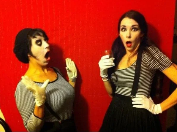 Two women with theatrical makeup and vintage-style striped shirts, wearing white gloves, standing against a red wall, making surprised expressions.
