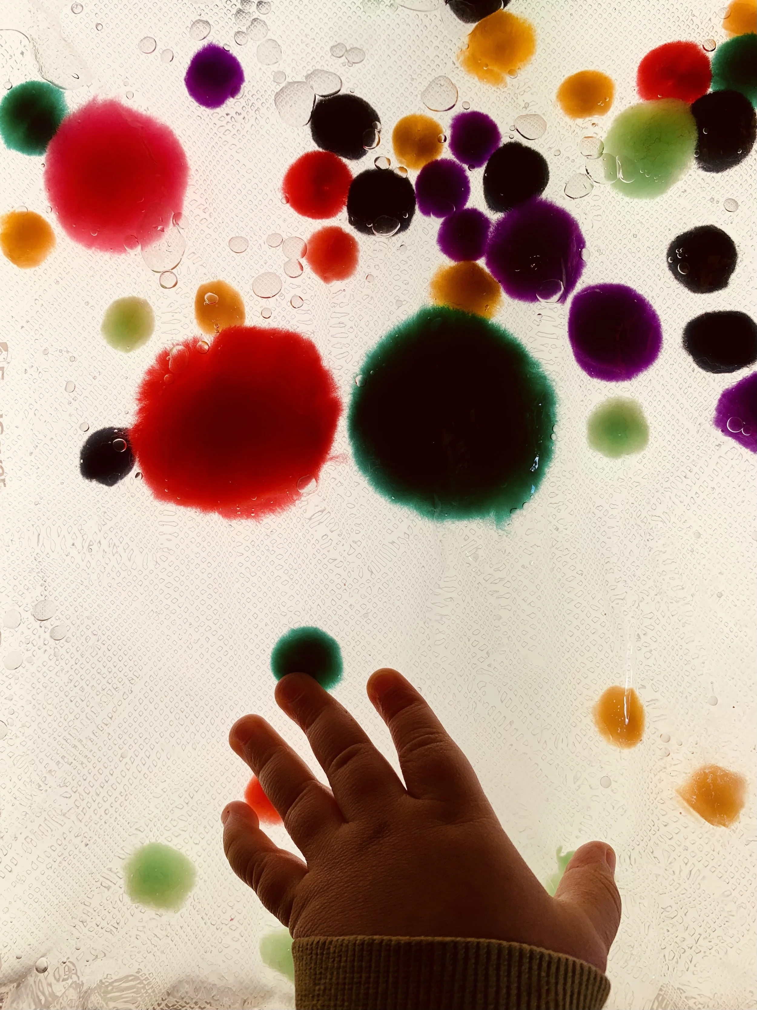 Colorful pom poms suspended in water on a light table with a hand reaching towards them, backlit to create a vivid, glowing effect.