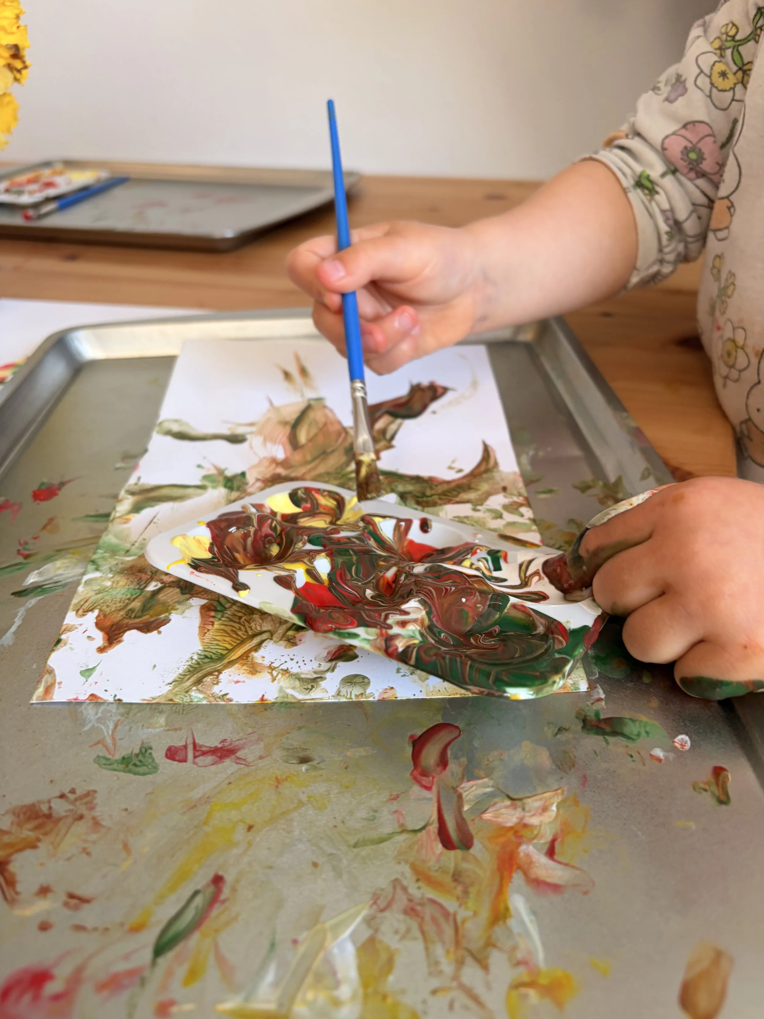 Child's hand holding a paintbrush, creating colorful abstract paint on a paper and splattered paints on a table.