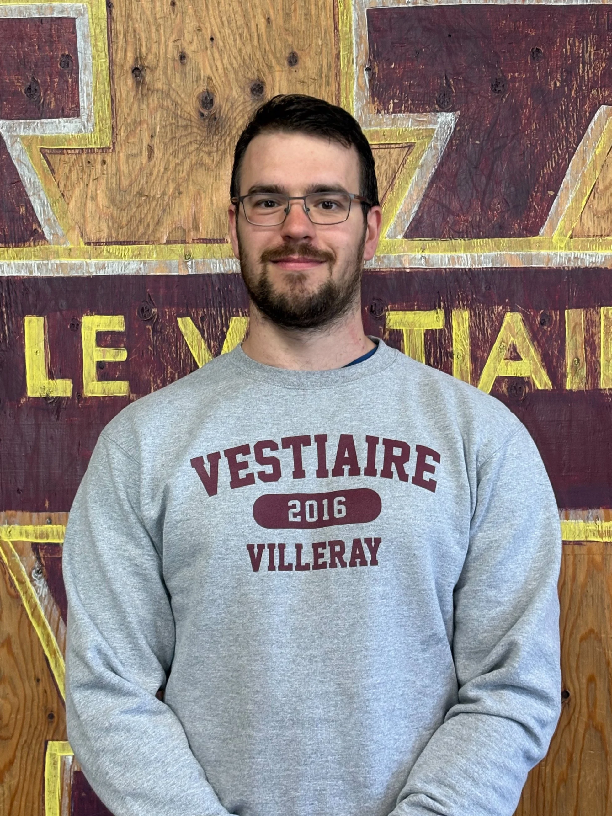 A man with glasses and a beard smiling, wearing a gray sweatshirt with maroon text, standing in front of a wooden background with painted letters.