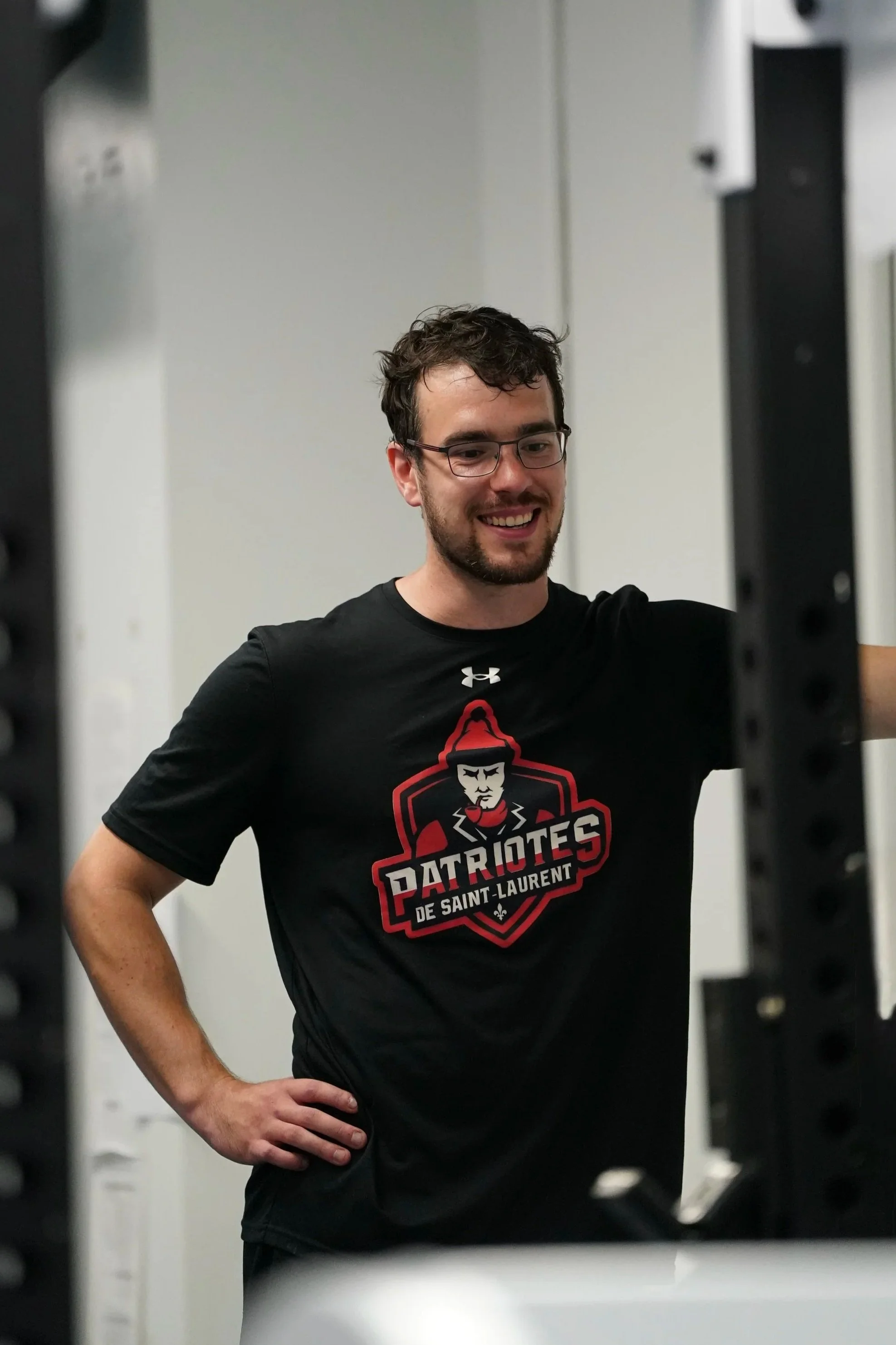 A young man with glasses and a beard smiling in a gym, wearing a black T-shirt with a sports team logo that reads "Patriotes de Saint-Laurent." He is standing with one hand on his hip, framed by gym equipment.