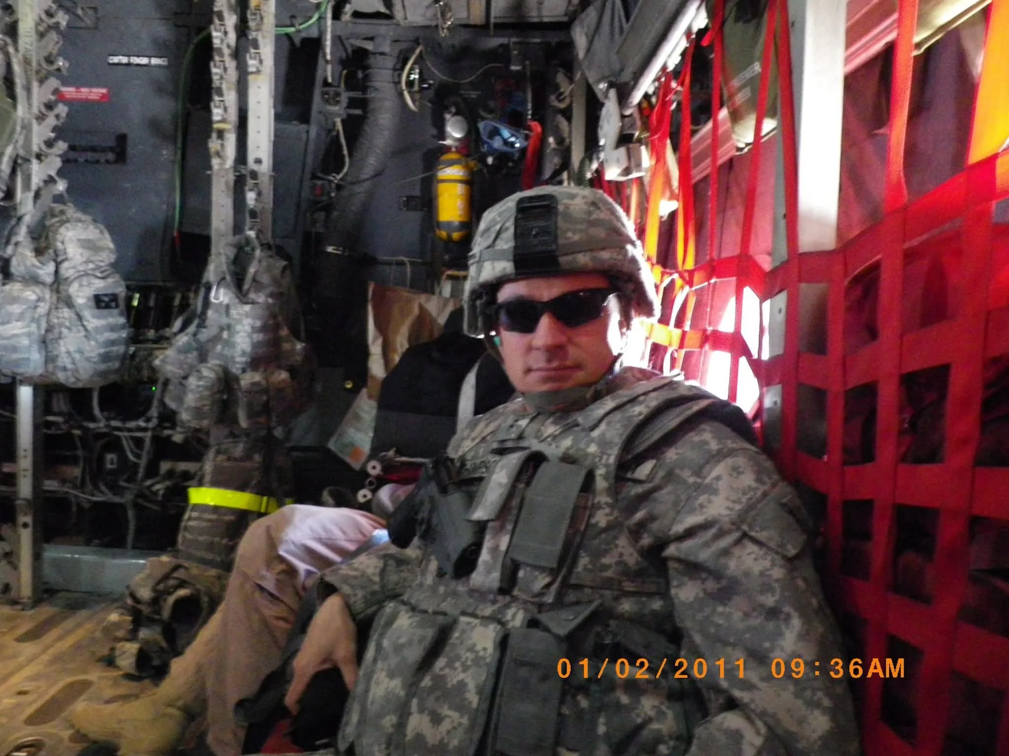 A man dressed in military camouflage uniform, wearing sunglasses and a helmet, appears to be sitting or taking a selfie inside an aircraft or helicopter. The background shows various equipment, wires, and a red metal structure. The photo has a timestamp at the bottom right corner indicating it was taken on January 2, 2011, at 9:36 AM.