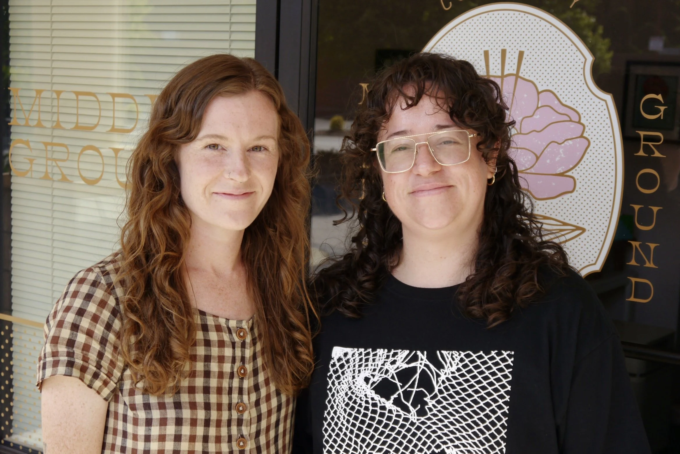 Two women with curly hair standing together outside a store with window blinds and a sign that reads 'MIDDLE GROUND' and a stylized flower logo.
