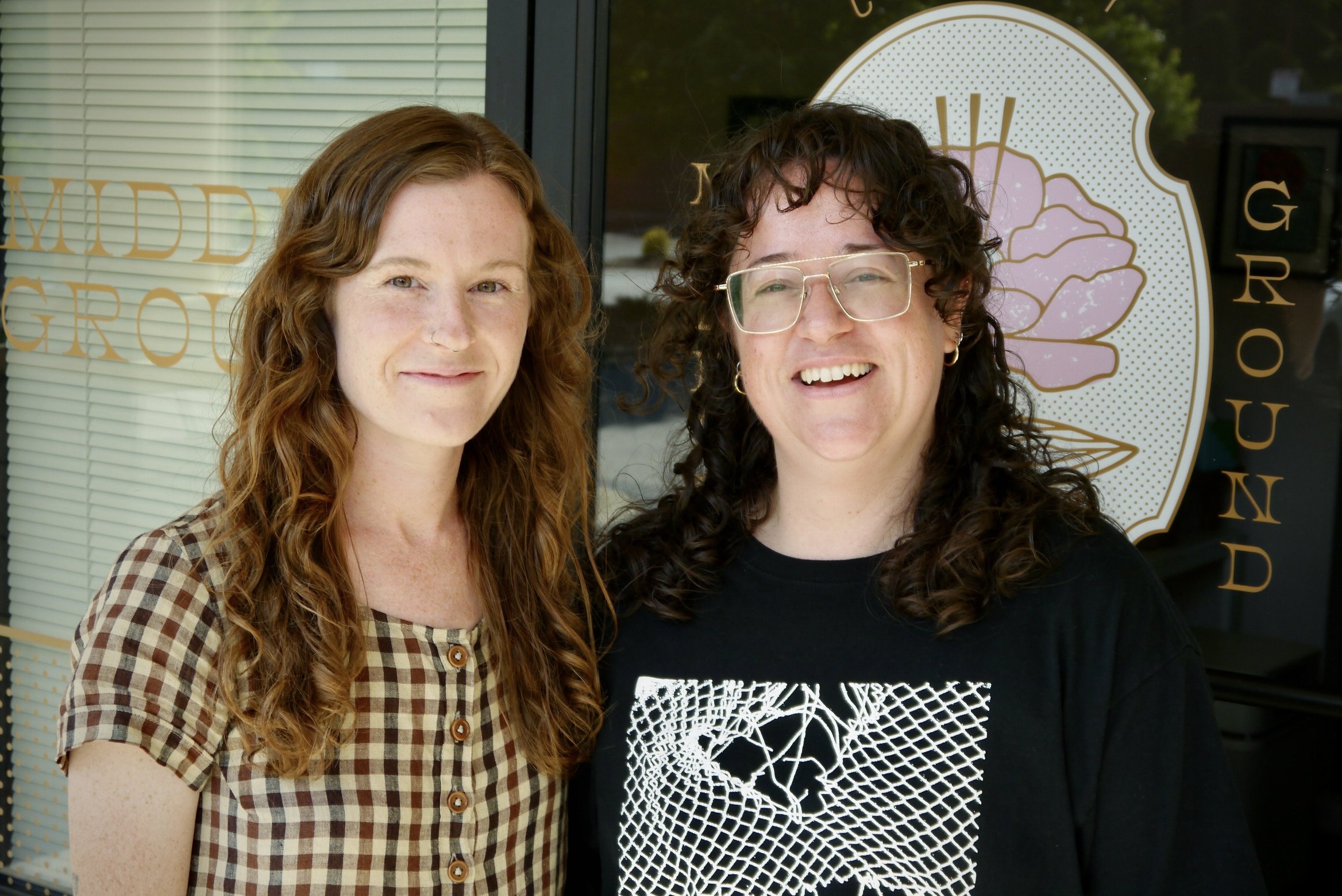 Two women standing outside, smiling at the camera. One with red curly hair wearing a checkered shirt, and the other with dark curly hair and glasses, wearing a black shirt with a geometric white design. Behind them is a storefront window with a pink graphic of a flower and the word 'GROUN' partially visible.