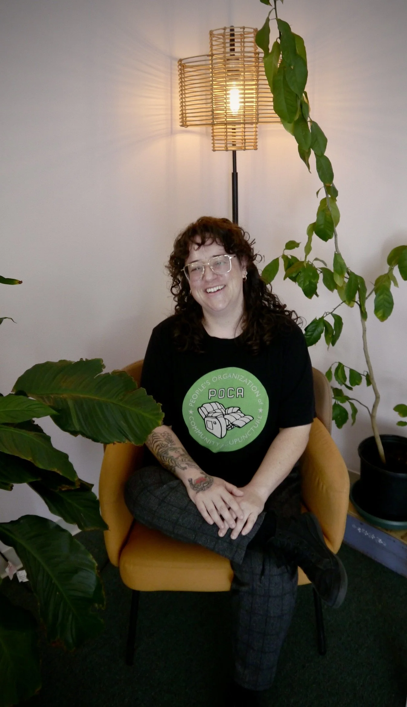 A smiling woman with curly brown hair, glasses, and tattoos on her arm, sitting in a yellow armchair surrounded by green plants, wearing a black t-shirt with a green logo. The background features a wall-mounted lamp.