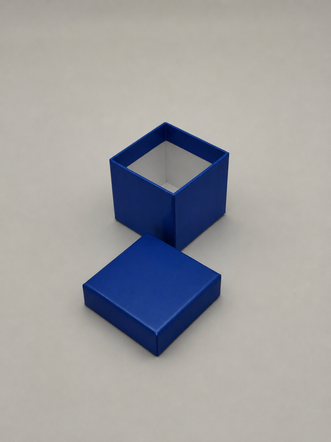 A small blue box with an open top and a matching square lid placed in front of it, on a plain light-colored surface.