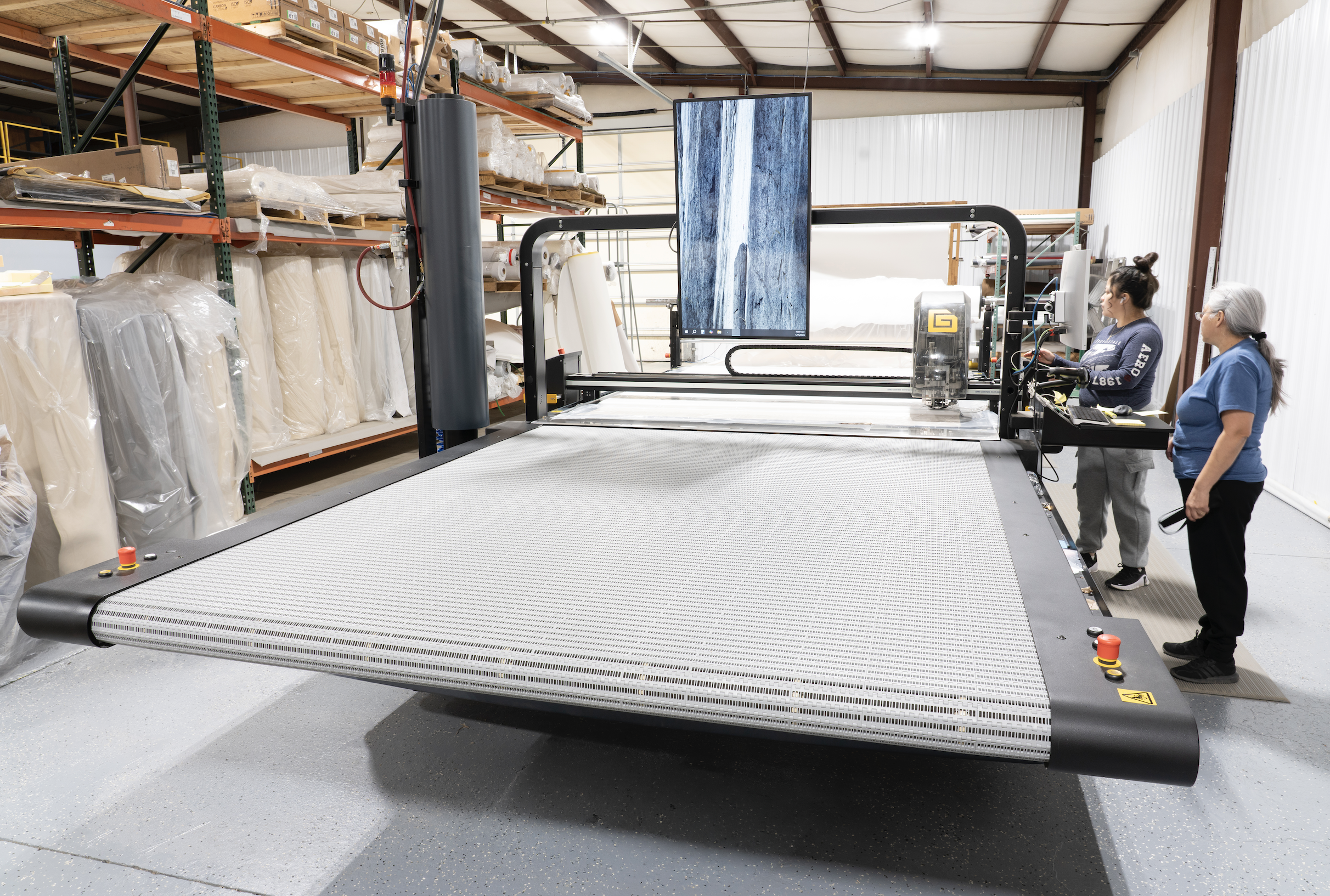 Two women observing a large industrial fabric printing machine inside a warehouse with storage shelves filled with fabric rolls.