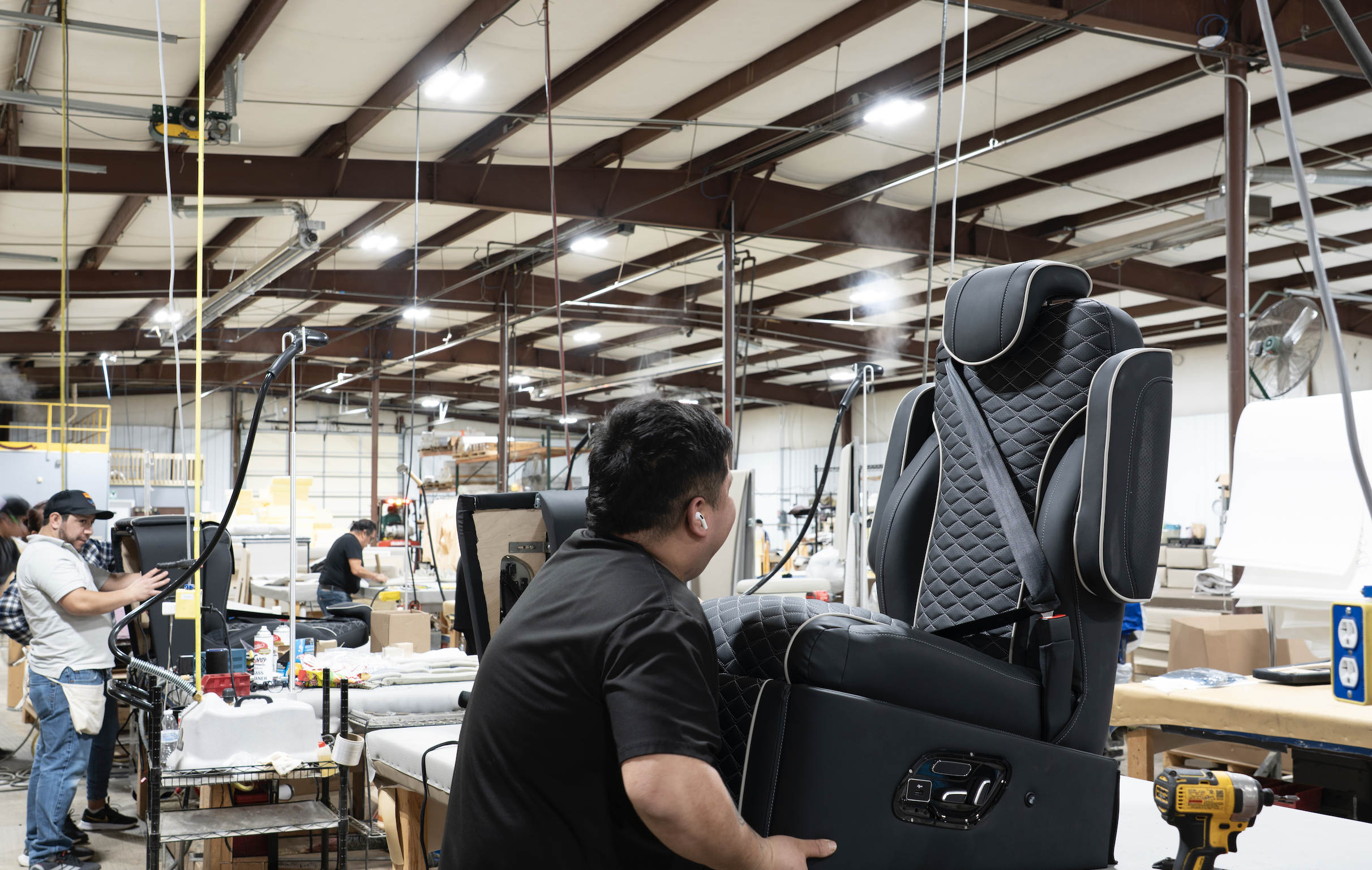 Workers assembling luxurious black leather chairs in a large factory with high ceilings and bright overhead lighting.