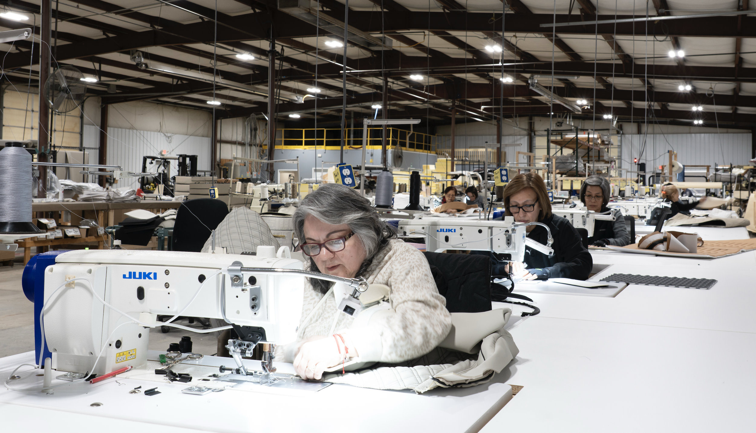 People working at sewing machines in a large factory with industrial shelving and supplies in the background.