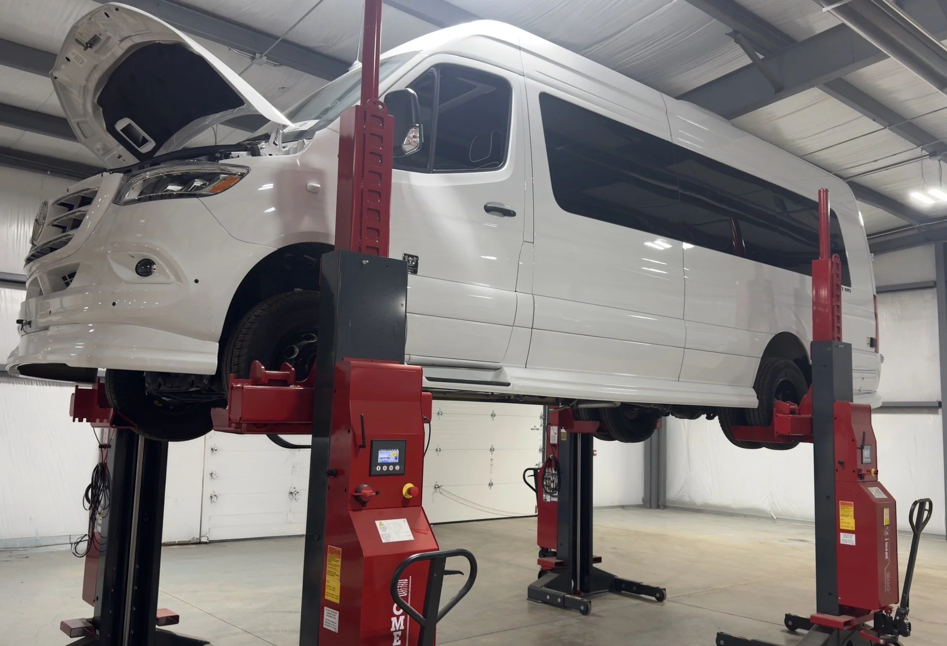 A white van is elevated on a vehicle lift inside a garage or service center. The van's hood is open, and the room has a high ceiling with metal beams and white walls.