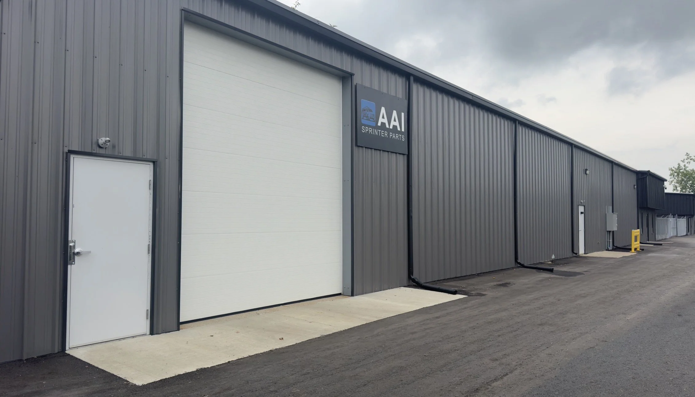 A large industrial building with gray metal siding, featuring a white roll-up garage door and a small white personnel door, with a sign reading 'AAI Sprinter Parts' on the building. The sky is overcast, and the ground is paved with asphalt.