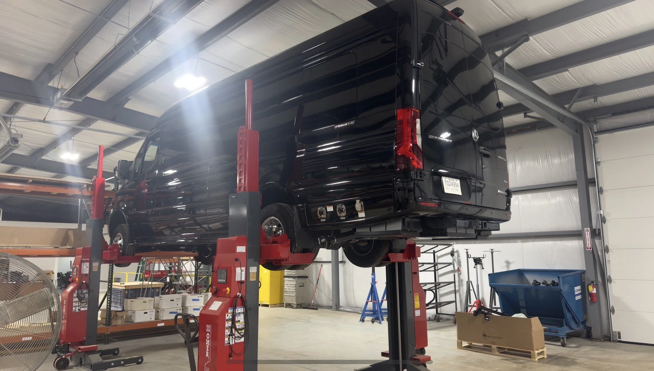 A black Mercedes-Benz Sprinter van elevated on a hydraulic lift inside a large indoor workshop with various tools, equipment, boxes, and a large industrial fan.