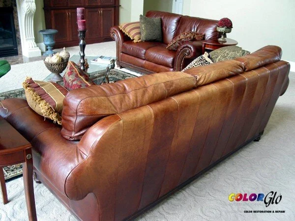 Living room with two brown leather couches, pillows, a glass coffee table, and decorative items. The room has a carpeted floor and a wooden cabinet in the background.