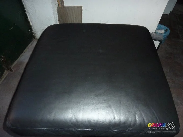 Close-up of a black leather ottoman in a room with dark flooring and a white wall.