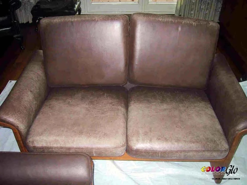 A vintage upholstered two-seater sofa with brown leather and fabric cushions, placed on a floor with some papers underneath.