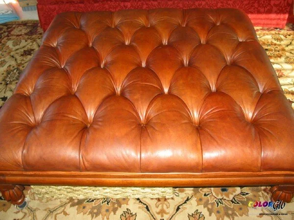 A close-up of a tufted leather ottoman with a wooden frame, placed on a patterned carpet.