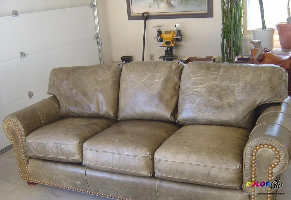 A worn brown leather couch with three cushions, showing visible scratches and signs of wear, in a room with a garage door, a shelf with a yellow tool, and houseplants.