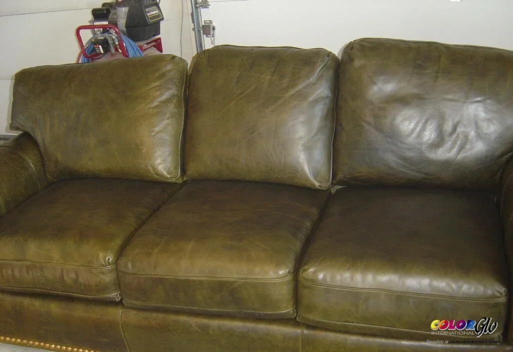 A three-seat greenish-brown leather sofa with visible wear and wrinkles, sitting in a room with a garage door in the background.