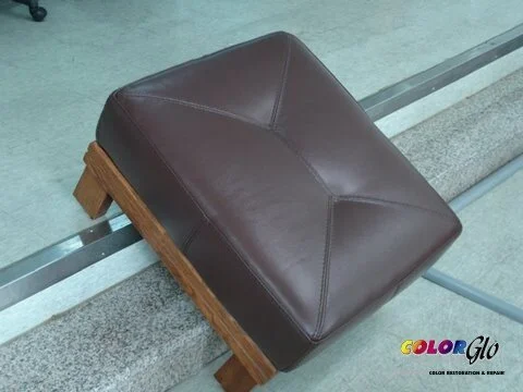 A brown leather cushioned stool with wooden legs placed on a concrete floor.