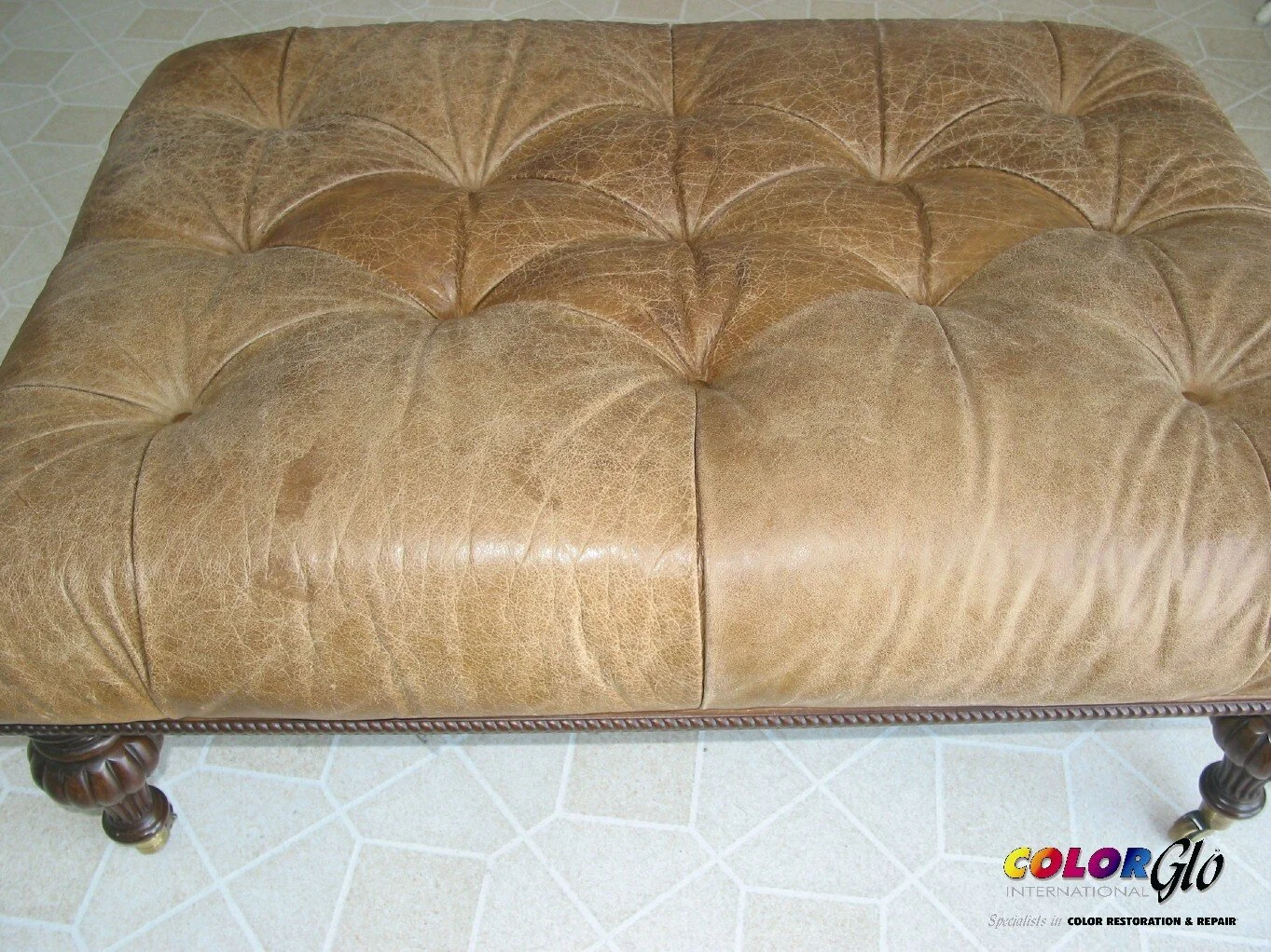 A rectangular vintage leather ottoman with tufted top and carved wooden legs, placed on a tiled floor.