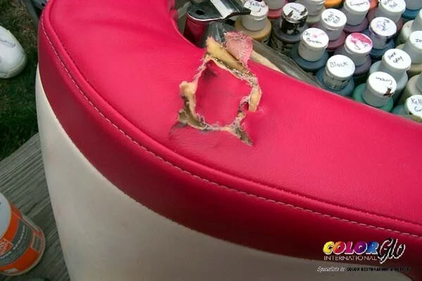 A red and white cushioned chair with a tear in the red upholstery revealing stuffing. In the background, there are numerous small bottles of paint or ink.