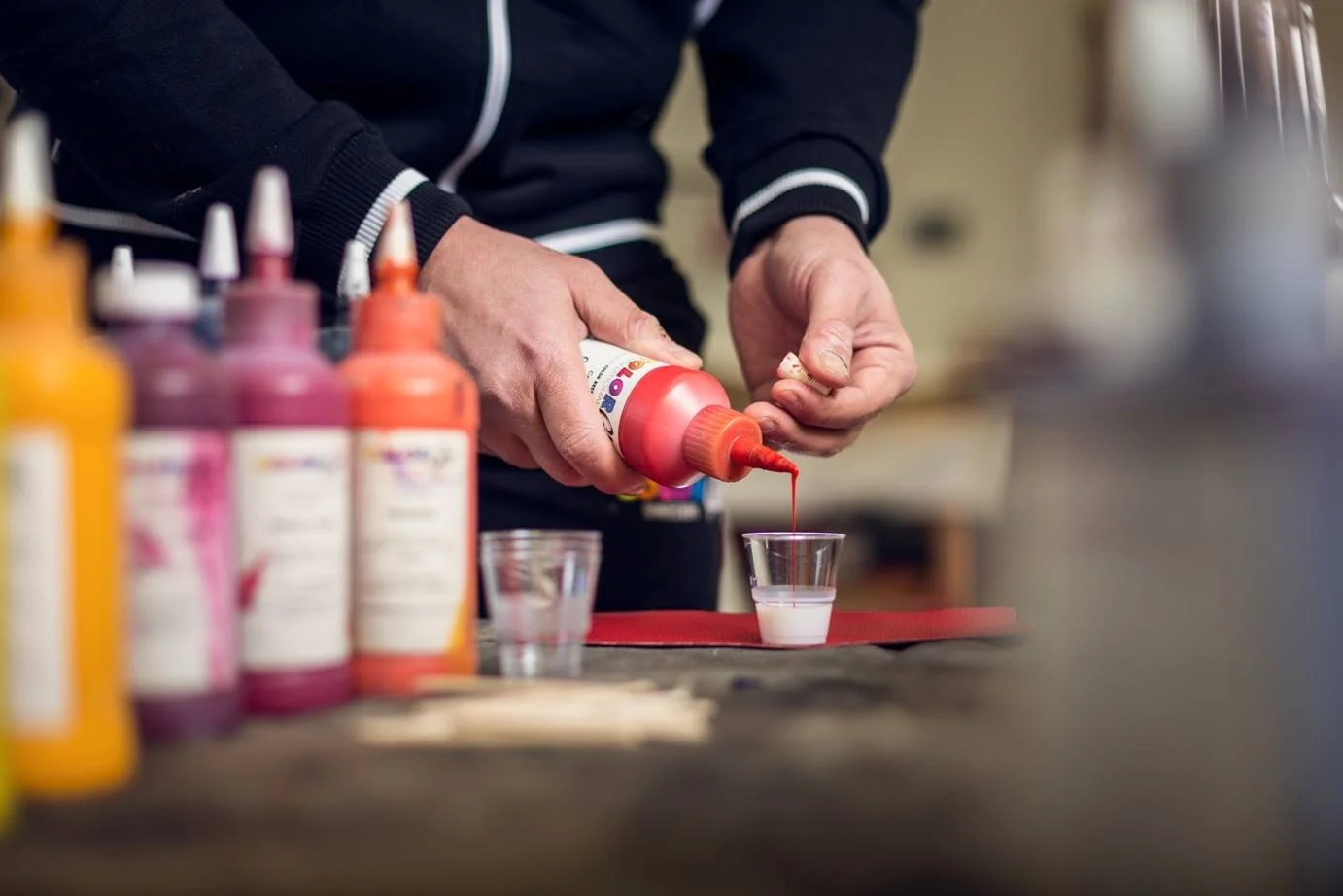 Person squeezing pink paint onto a small clear cup, with various bottles of paint in the background.