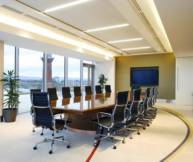 Modern conference room with a large wooden table, black leather chairs, floor-to-ceiling windows, potted plants, and a large flat-screen TV