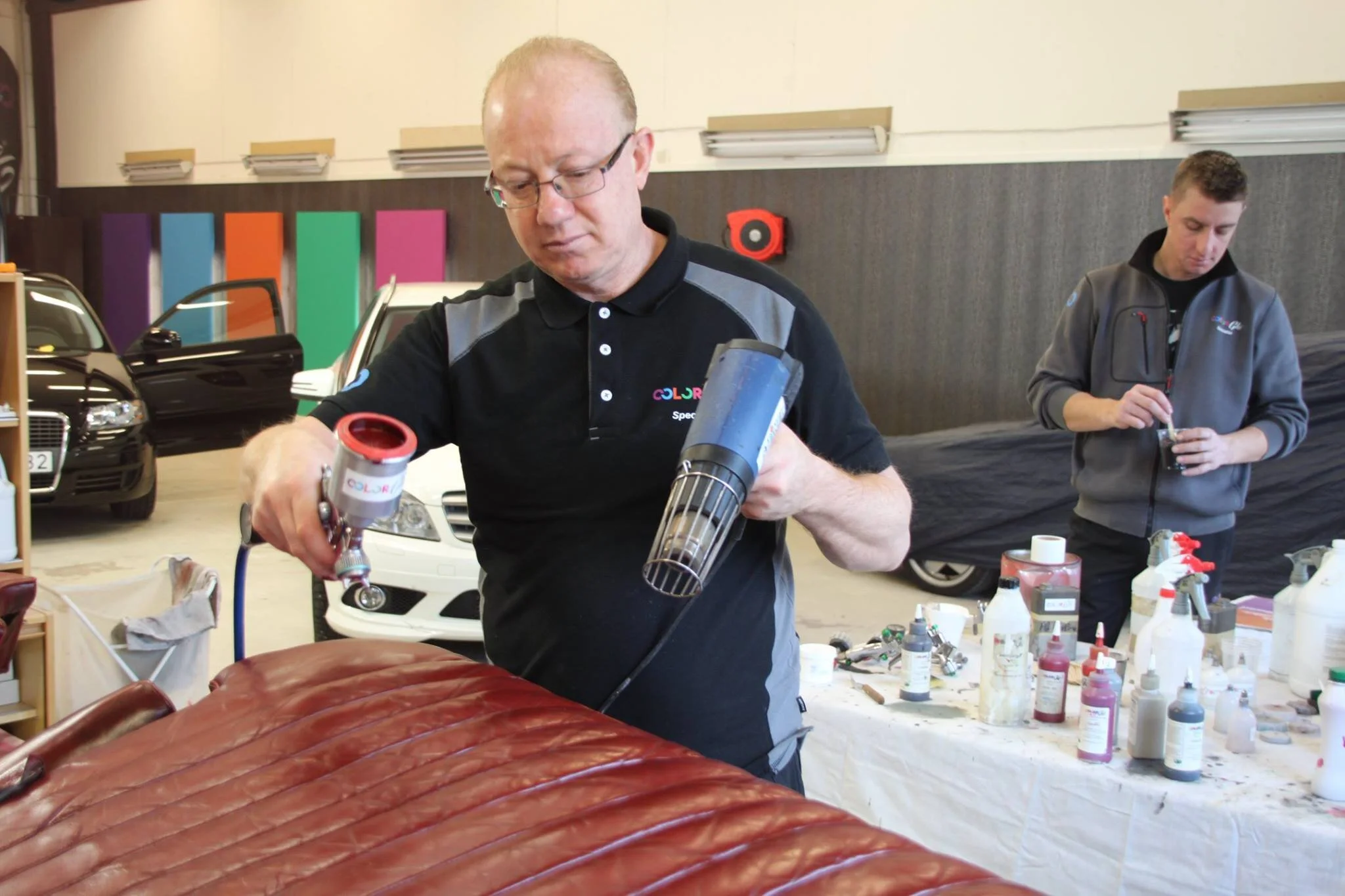 Two men working on a piece of furniture or leather sofa at a workshop or store, with various paints and supplies on the table in front of them.