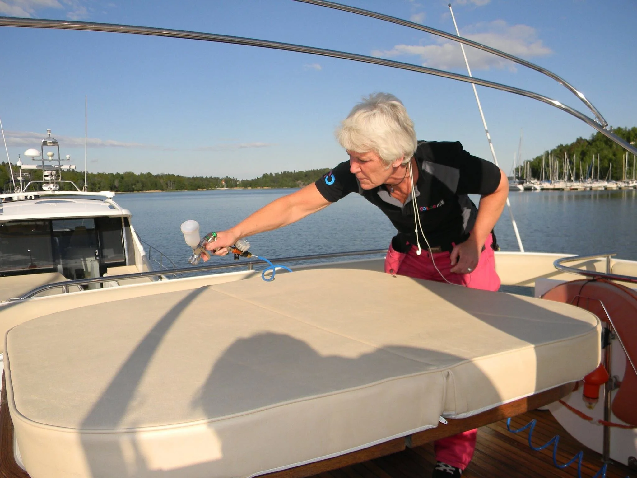 An elderly woman with short white hair wearing a black polo shirt with logos and pink pants working on a sailboat, adjusting equipment on the deck. The scene is set outdoors on a marina with other boats and sailboats docked in the background, under a clear blue sky.