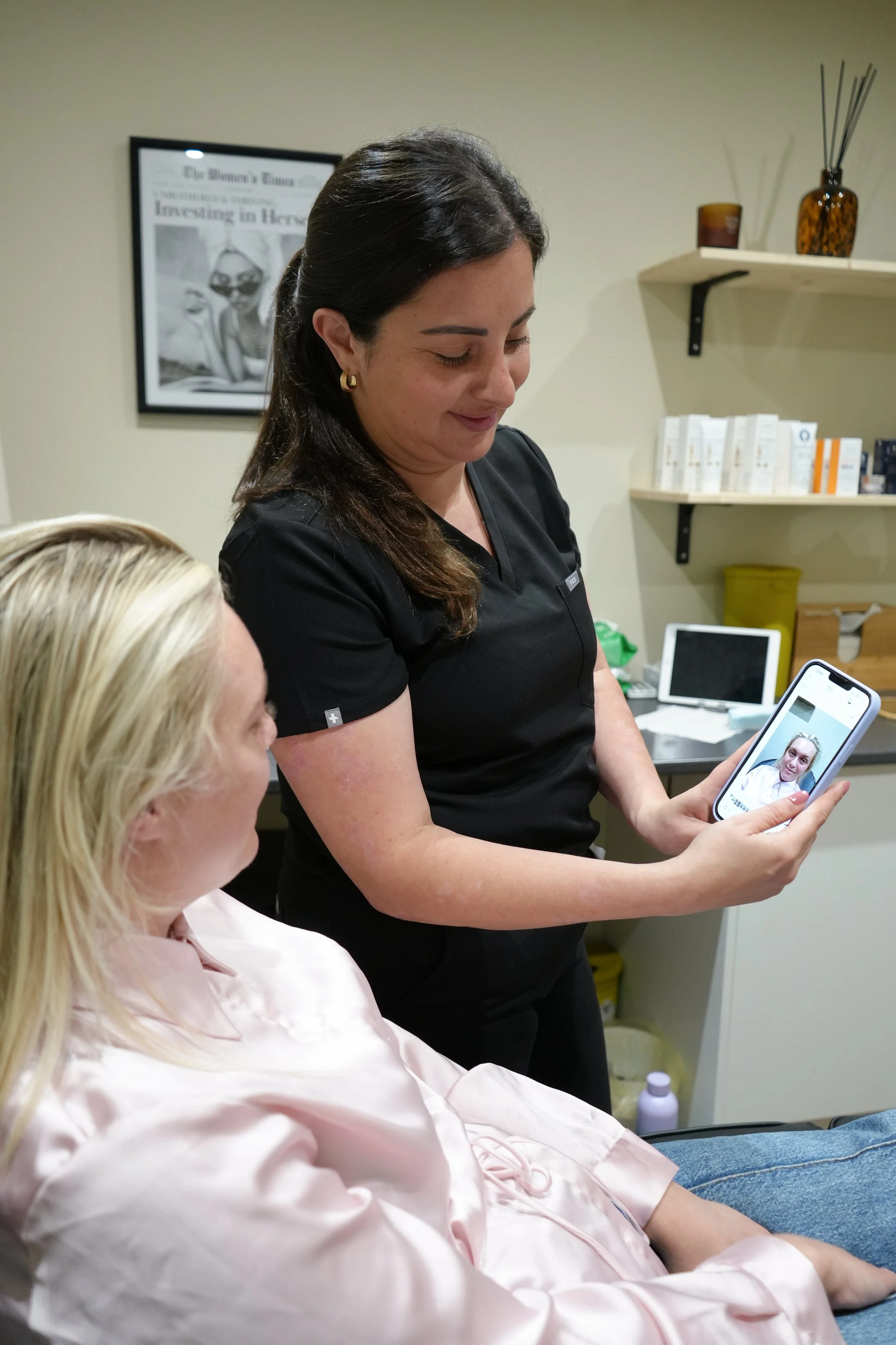 A woman with dark hair and earrings looking at her smartphone. A woman with blonde hair wearing a pink shirt is seated in front of her. The setting appears to be a medical or professional environment with shelves, books, and a framed newspaper clipping on the wall.