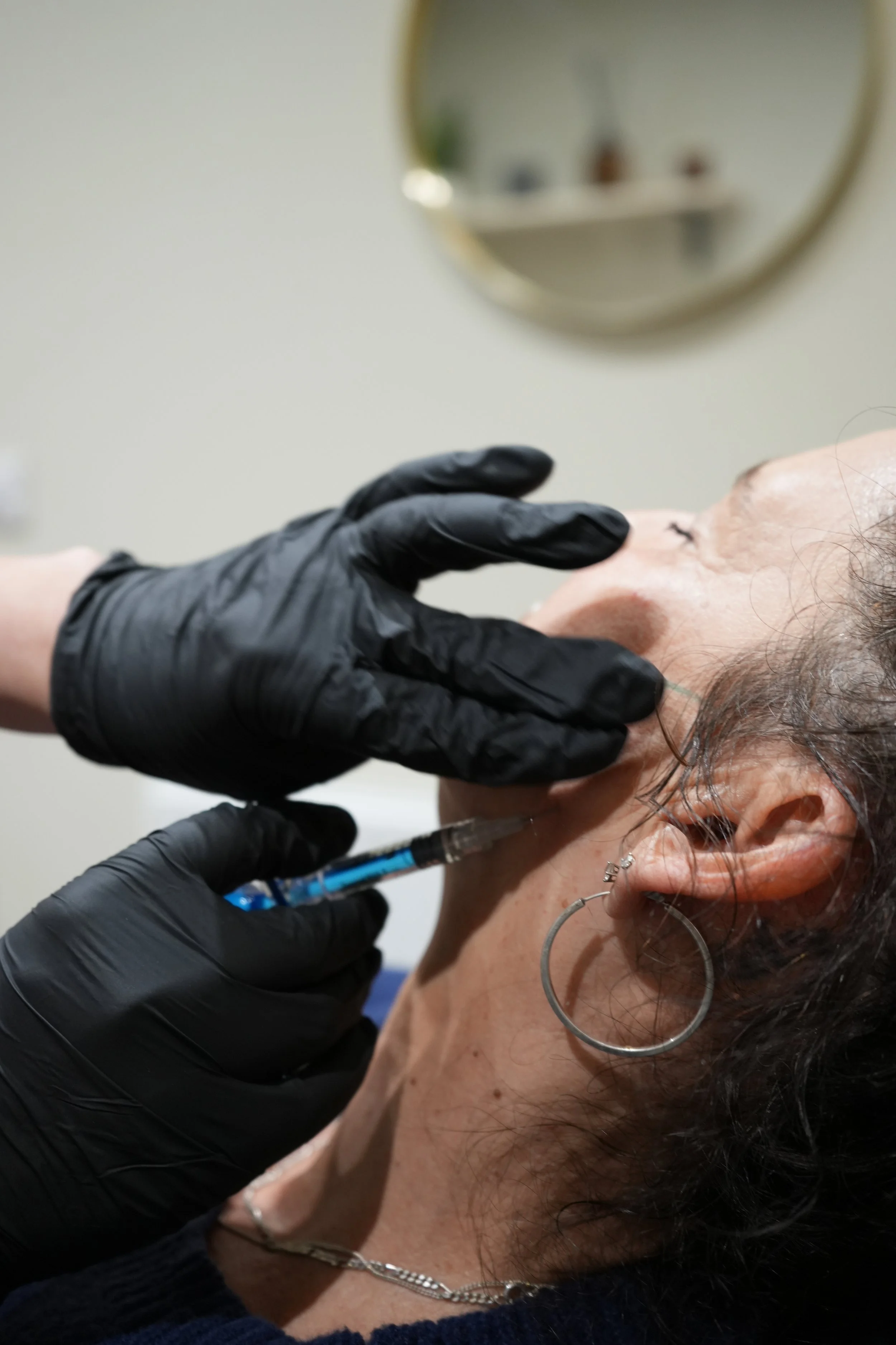 A healthcare professional wearing black gloves administering an injection into a person's neck area with a syringe. The person receiving the injection has earrings and a necklace, with a mirror and some blurred objects in the background.
