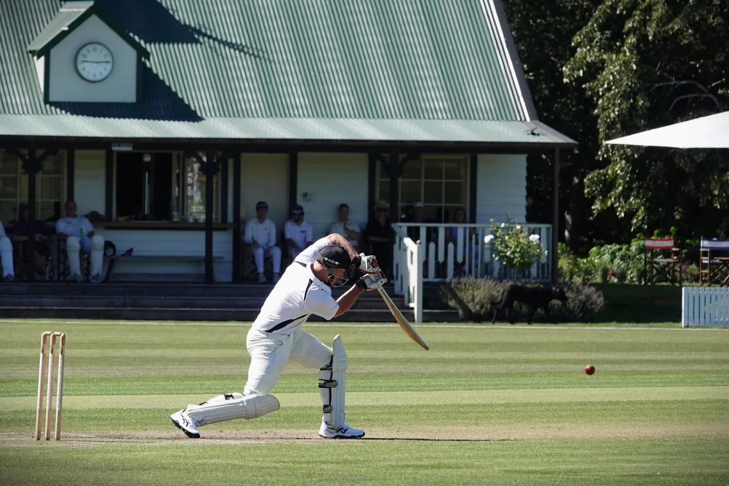 Cricket player batting on the field with spectators watching from a porch in the background.