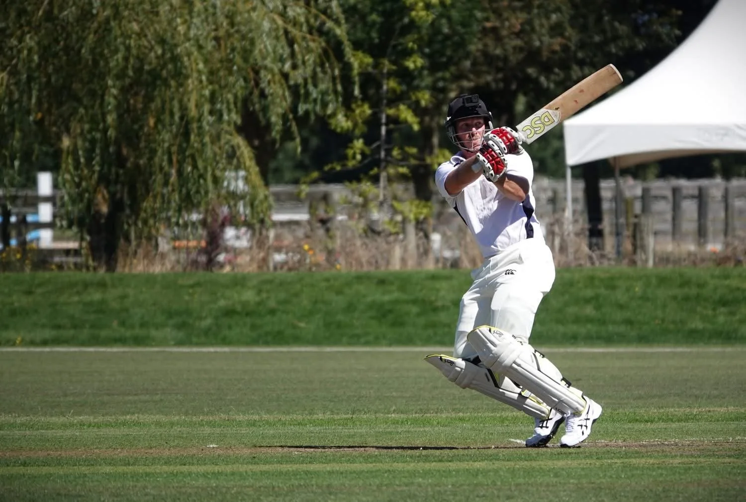 A cricket player in white uniform and protective gear swings a bat on a grassy field.