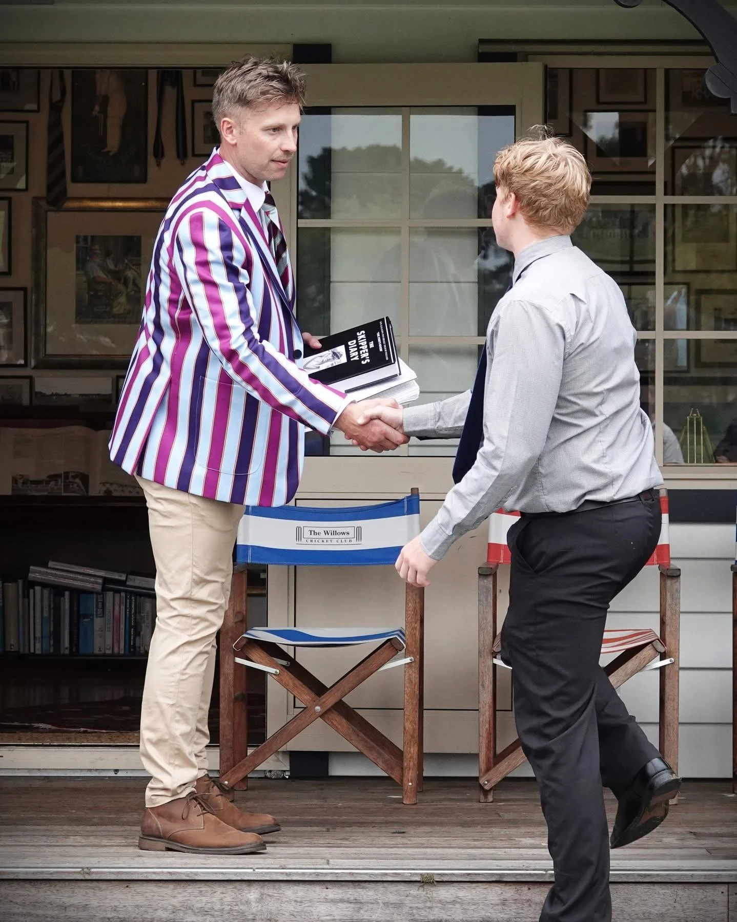 Two men shaking hands outdoors, one wearing a colorful striped blazer and the other in a gray shirt and black pants. The man in the blazer is holding a book titled 'Supery's Diary'. There are two wooden chairs and a small bookshelf behind them.