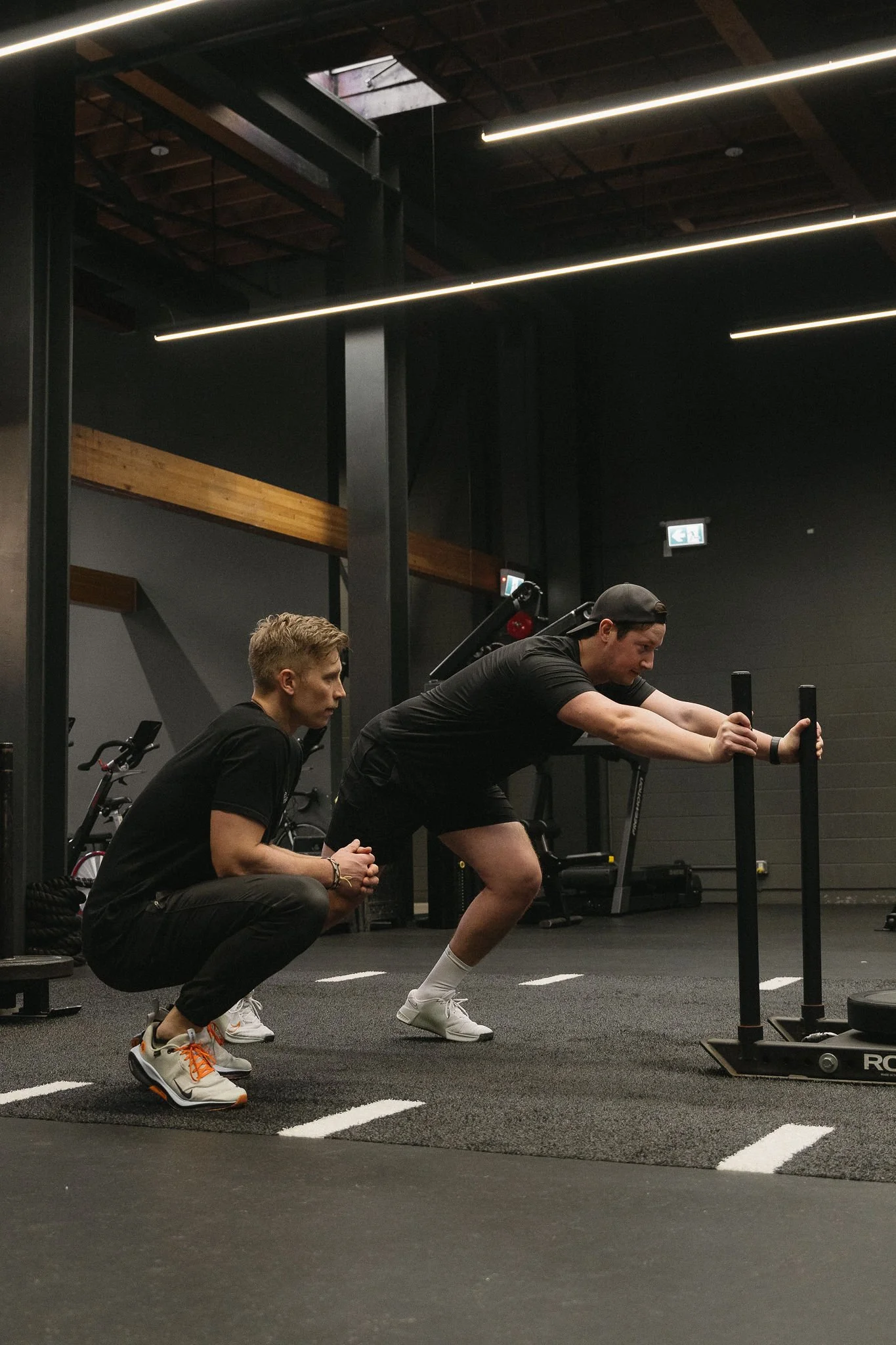 Two men working out at gym, one squatting with a barbell, the other squatting beside him, and a third man crouching in front of them observing.