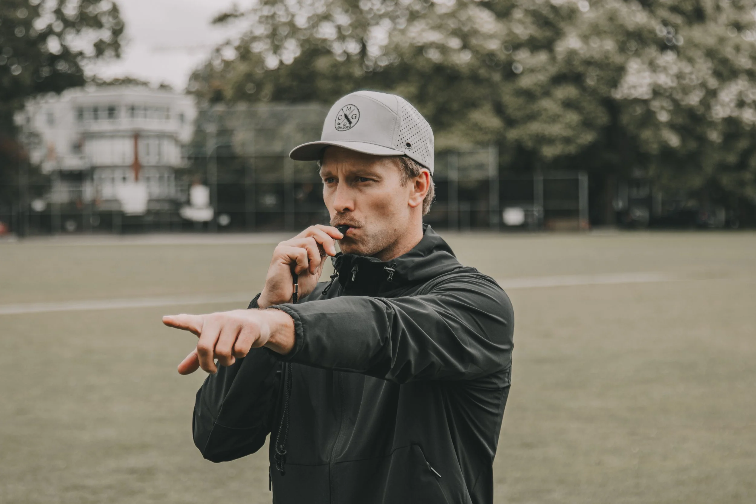Man in black jacket and gray cap with logo, outdoors on a field, appearing to stretch or warm up.