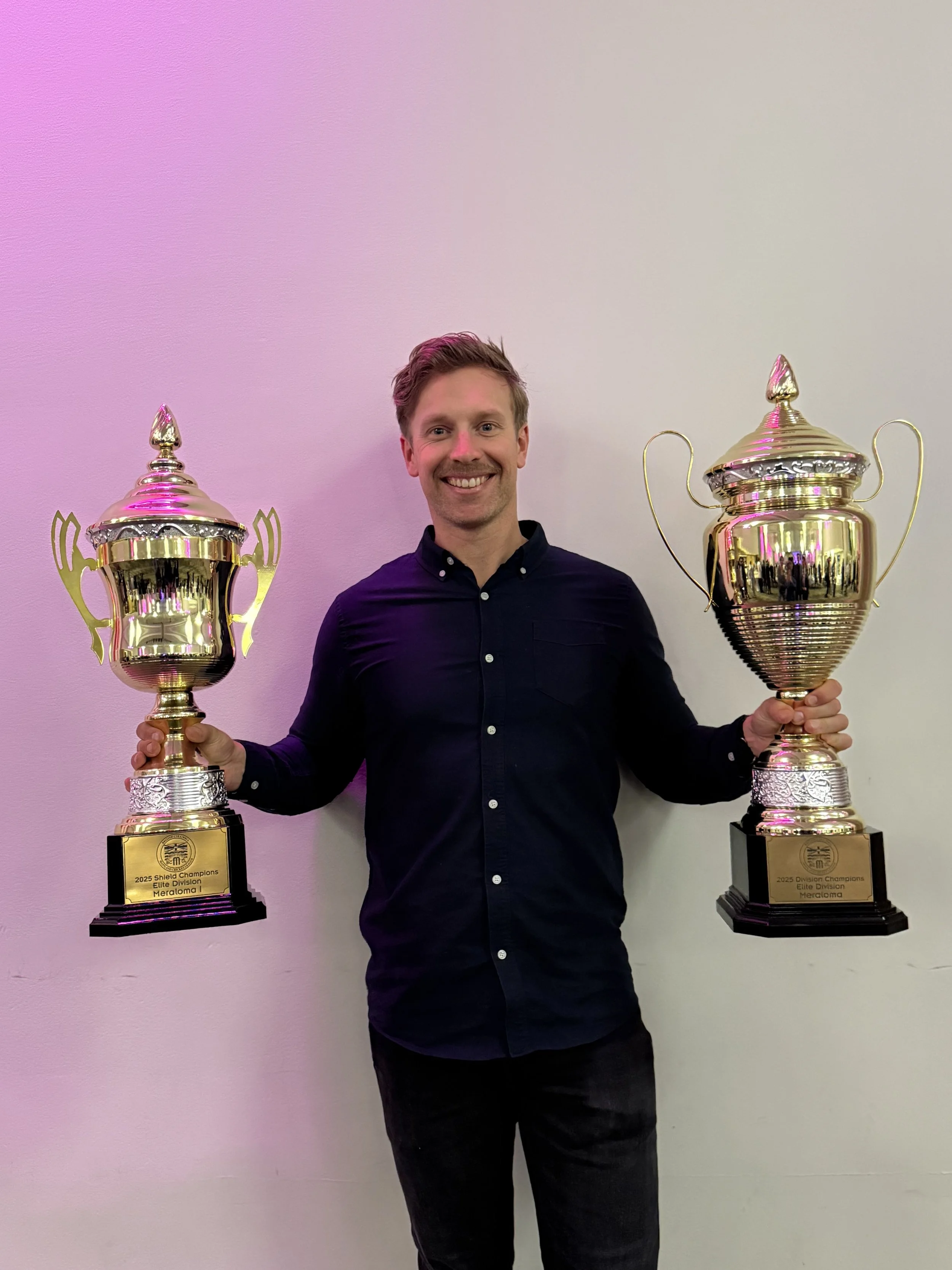 A man in a black shirt holding two gold trophies, standing against a plain white wall.