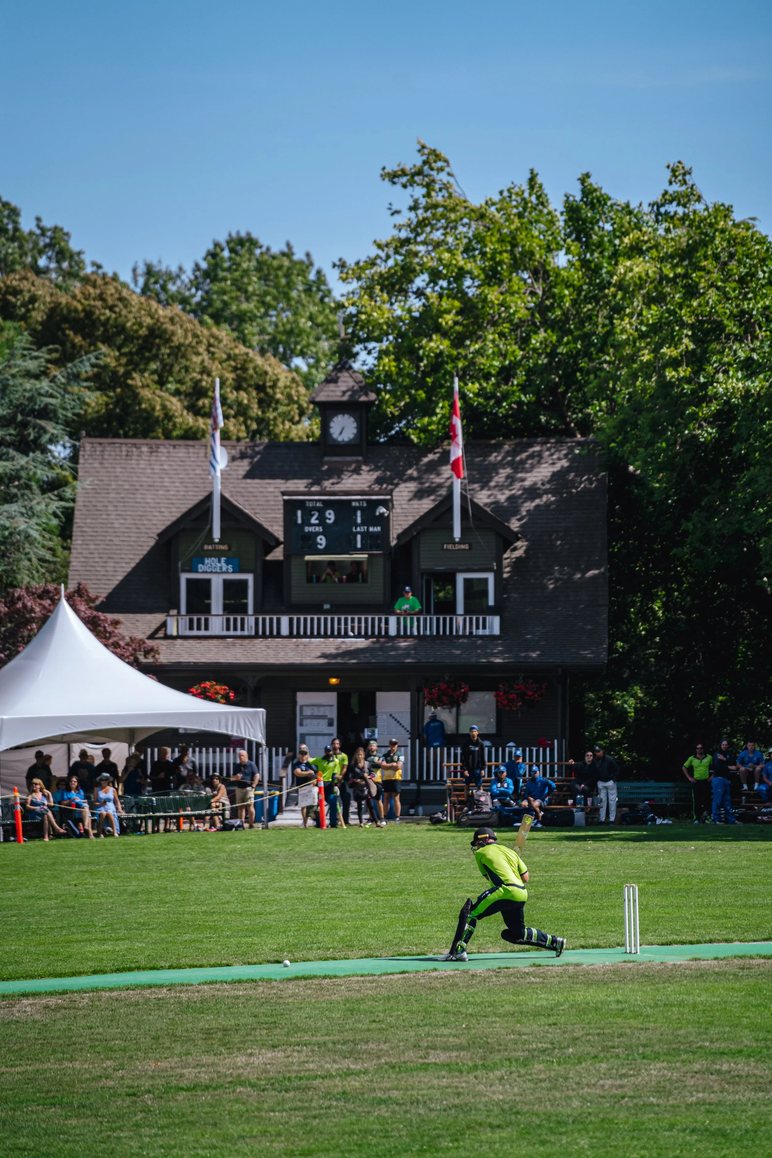 Cricket match scene with a bowler in bright yellow and black uniform bowling on a well-maintained grass field. Spectators are seated under tents and on seats, watching the game. In the background, a scoreboard displays scores, and two flags are flyin