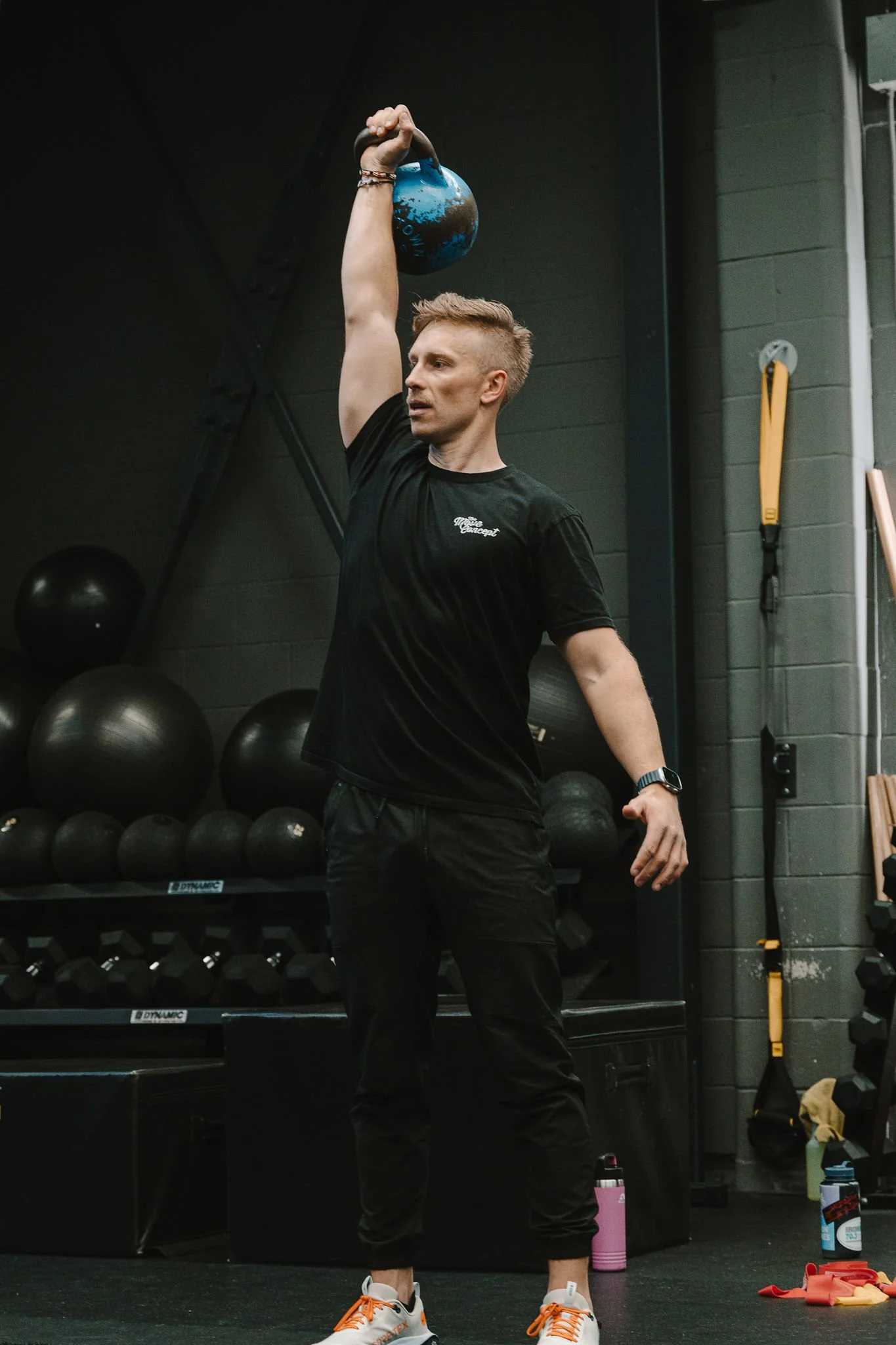 A man in black workout clothes lifting a blue kettlebell overhead in a gym with black walls and exercise equipment in the background.