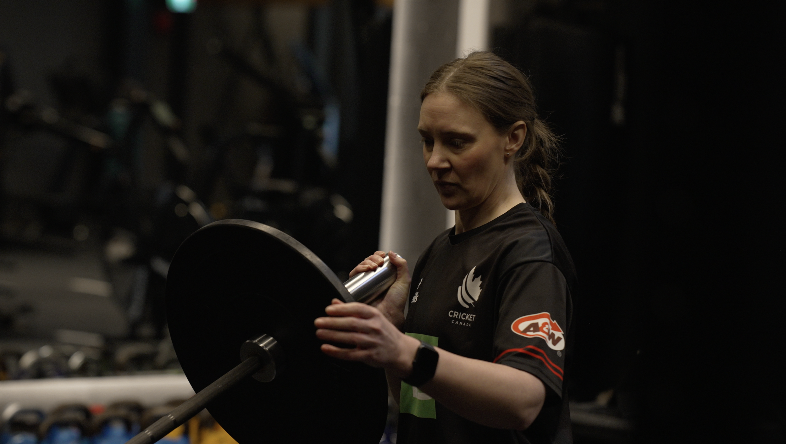 A woman with brown hair in a ponytail wearing a black sports shirt with a Cricket Canada logo and an A&W logo, holding a dumbbell in a gym.
