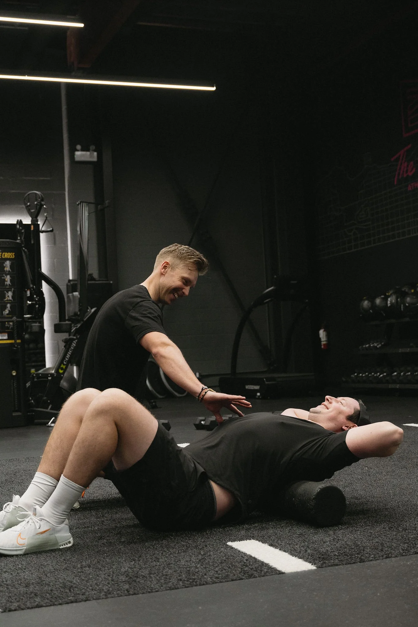 Two men working out in a gym, with one lying on a foam roller and the other assisting with a stretch. The gym has black walls, workout equipment, and a black floor.