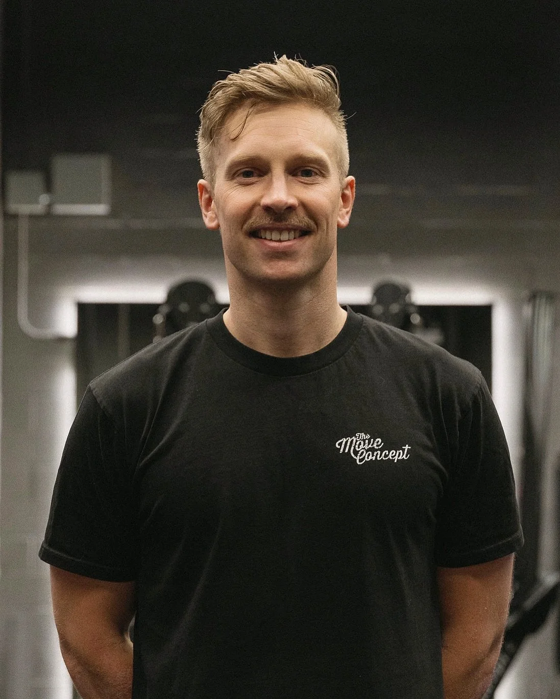 A young man with short light brown hair and a mustache smiling at the camera, standing in a gym wearing a black T-shirt that says 'The Move Concept'.