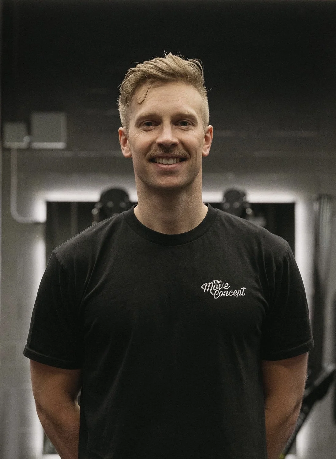 A smiling man with short blond hair in a black t-shirt stands in a gym, with gym equipment and a dark ceiling in the background.