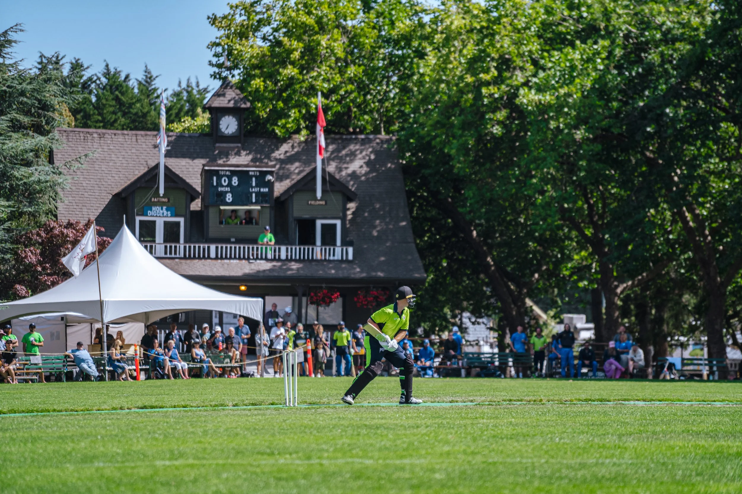 Cricket match with a player batting on a green field, crowd watching from the stands, and a scorer's booth on a large building behind, surrounded by trees on a sunny day.